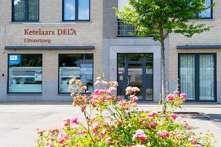 Gevel van een gebouw met de tekst “Ketelaars DELA Uitvaartzorg”, met ramen, voordeur en bloemen op de voorgrond langs de straat.